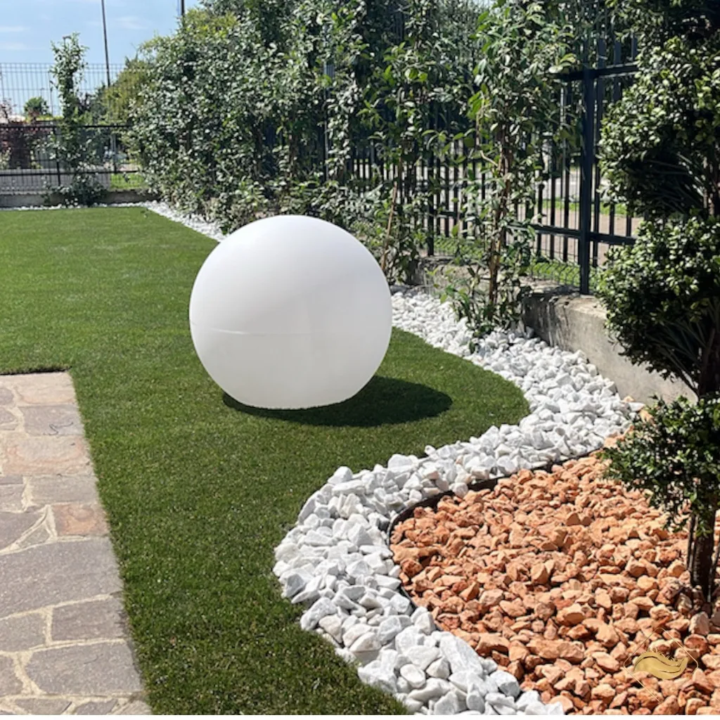 Sfera luminosa decorativa su prato sintetico con aiuola in ghiaia bianca e rossa, perfetta per illuminare e valorizzare il giardino con stile.